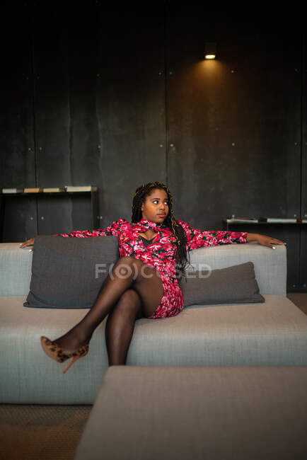Afro-americano feminino em vestido rosa brilhante e saltos altos sentado no sofá no lobby do hotel — Fotografia de Stock