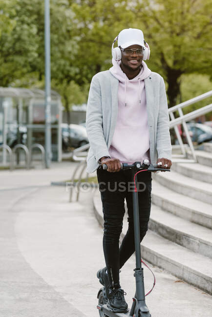 Corpo inteiro de afro-americano masculino em fones de ouvido sem fio montando scooter elétrico perto de escadaria na cidade — Fotografia de Stock