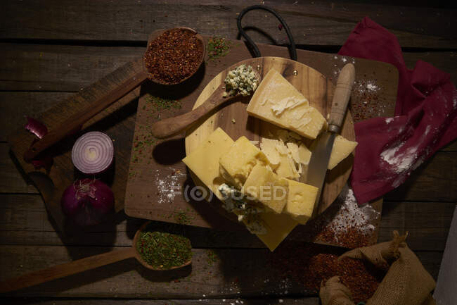 Сверху нарезанные кусочки вкусного сыра пармезан на разделочной доске помещены среди ароматических сушеных специй и свежего лука во время приготовления пищи на деревянном столе — стоковое фото