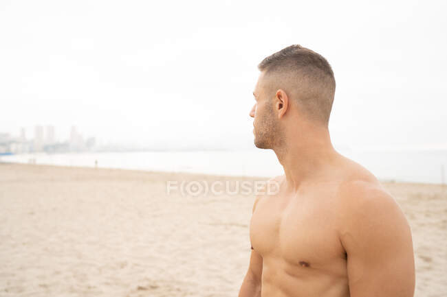 Determined male athlete with muscular naked torso standing on seashore in summer and looking away — Stock Photo