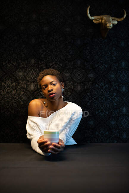 Donna afroamericana serena seduta a tavola con tazza di bevanda rinfrescante e guardando la fotocamera nella stanza buia — Foto stock