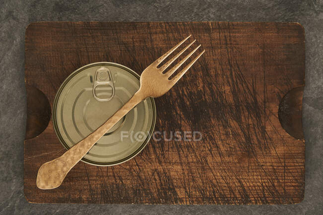From above scratched chopping board with fork and sealed can with preserved food on rustic lumber table — Stock Photo