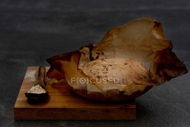 Composição com massa de pão rústica cozida na hora pão redondo em pergaminho colocado em tábua de madeira com colher e farinha de trigo — Fotografia de Stock