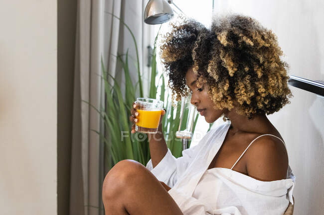Seitenansicht einer Afroamerikanerin in weißer Nachtwäsche, die zu Hause frischen Orangensaft aus dem Glas trinkt — Stockfoto