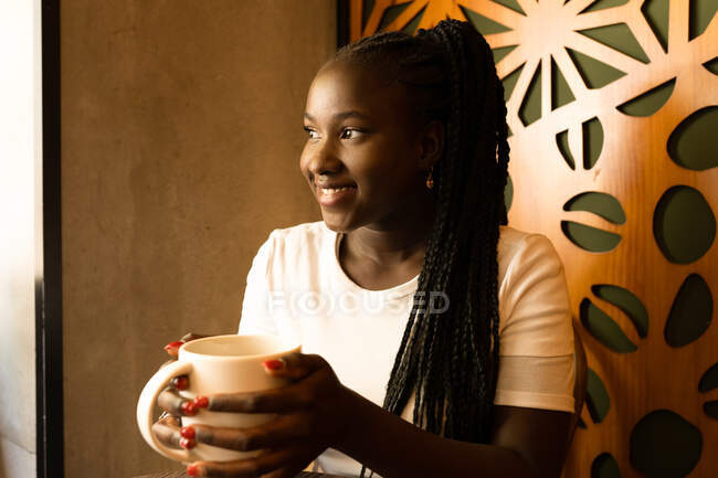 Positive black female with braids and mug of aromatic drink chilling in cafe and looking away — Stock Photo