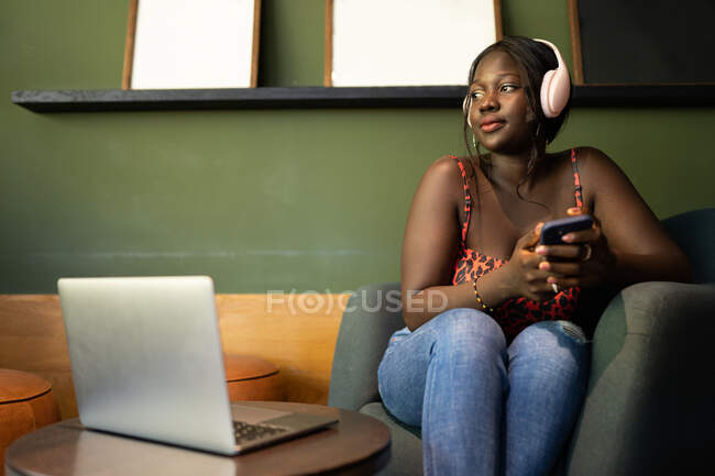 Donna nera ascoltare musica dal suo cellulare in una caffetteria mentre in piedi accanto a un computer portatile — Foto stock