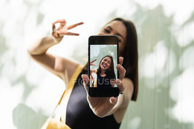 Femme joyeuse avec la langue en prenant soi-même tourné sur smartphone tout en montrant geste de paix et en s'amusant en ville en été — Photo de stock