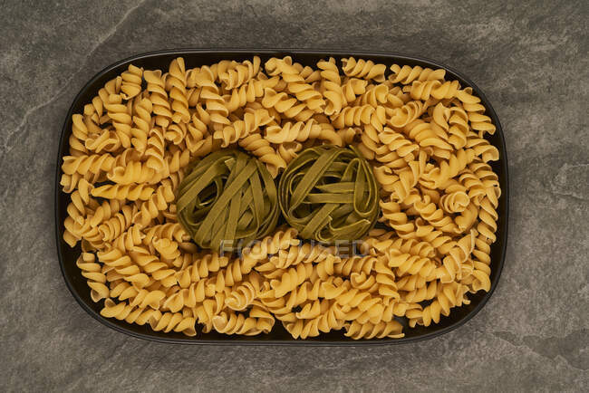 Top view of tray filled with raw fusilli and tagliatelle pasta and placed on gray table — Stock Photo