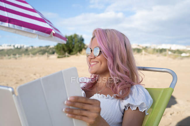 Mujer freelancer feliz sentado en la tumbona y trabajando en la tableta en la playa de arena cerca del mar en verano - foto de stock