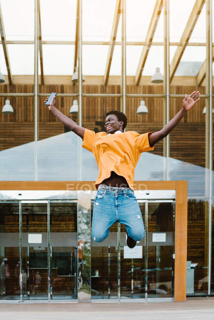 Basso angolo corpo pieno di maschio afroamericano con cuffie sul collo saltando in alto con le braccia tese — Foto stock