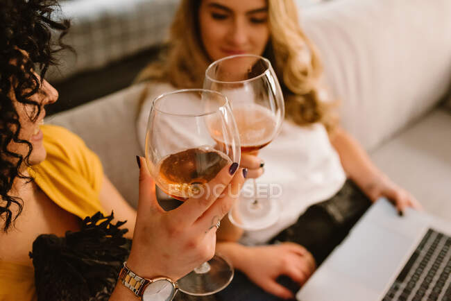 Crop content lesbian couple in casual wear clinking glasses with white wine while sitting on cozy couch and using netbook — Stock Photo