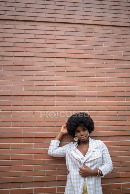 Young curly haired African American female model in checkered shirt and earrings leaning against red brick wall and looking at camera — Stock Photo