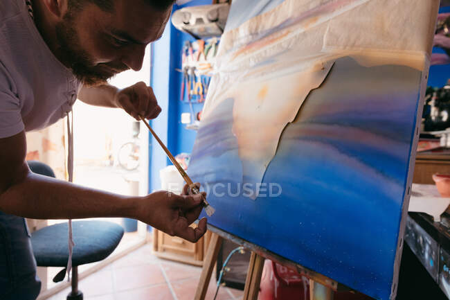 Hombre barbudo pintando puntos con pigmento blanco sobre lienzo con cuadro abstracto durante el trabajo en taller creativo - foto de stock