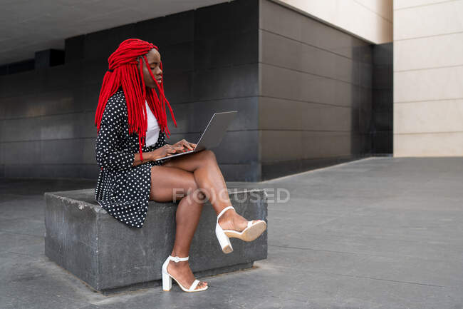 Vista lateral cuerpo completo de afroamericana enfocada freelancer escribiendo en netbook mientras trabaja remotamente al aire libre - foto de stock
