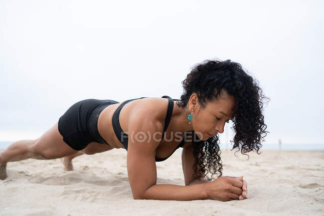 Vue latérale de l'athlète féminine en forme faisant de l'exercice de planche pendant l'entraînement sur le rivage sablonneux — Photo de stock