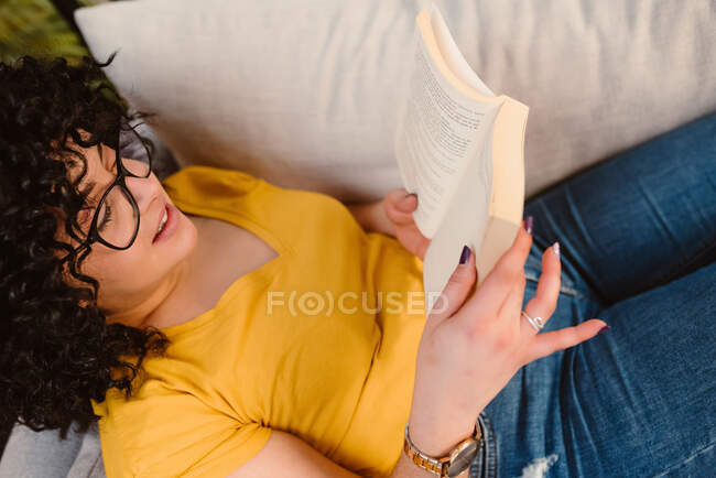 Desde arriba de la cosecha joven hembra en gafas con pantalones vaqueros y camiseta libro de lectura mientras está acostado en el sofá - foto de stock
