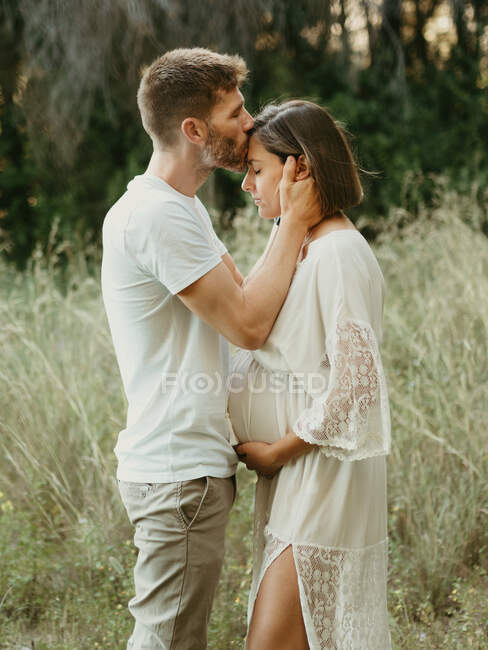 Vista lateral do homem macio beijando fêmea grávida na testa, enquanto em pé em campo na natureza — Fotografia de Stock