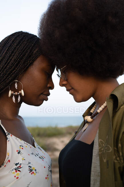 Side view of young African American female best friends in stylish outfits and bijouterie standing close with eyes closed on seashore in summer day — Stock Photo