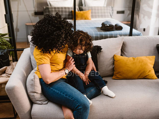 Madre con el pelo rizado abrazando niño adorable mientras se sienta en un cómodo sofá juntos en la sala de estar - foto de stock