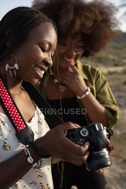 Jovens amigas afro-americanas alegres em roupas elegantes de verão e acessórios verificando fotos na câmera fotográfica enquanto passam férias de verão juntas no campo — Fotografia de Stock