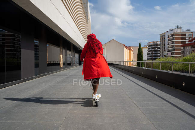 Visão traseira corpo inteiro da fêmea afro-americana com tranças brilhantes montando scooter elétrico ao longo do edifício da torre no tempo livre — Fotografia de Stock