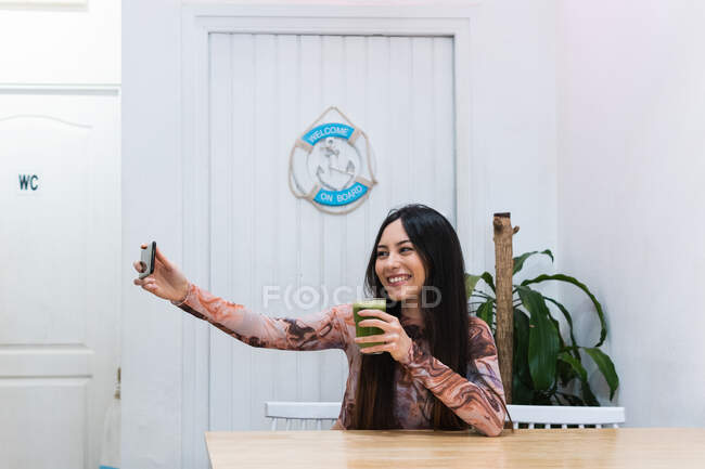 Позитивна жінка сидить за столом з коктейлем, а сама користується мобільним телефоном під час холоду на вихідних у барі. — стокове фото
