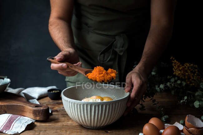 Cocinero macho irreconocible recortado usando cuchara de madera para mezclar puré de calabaza con huevos y harina en un tazón mientras prepara pastel en la mesa de madera cerca de la tabla de cortar y la toalla - foto de stock