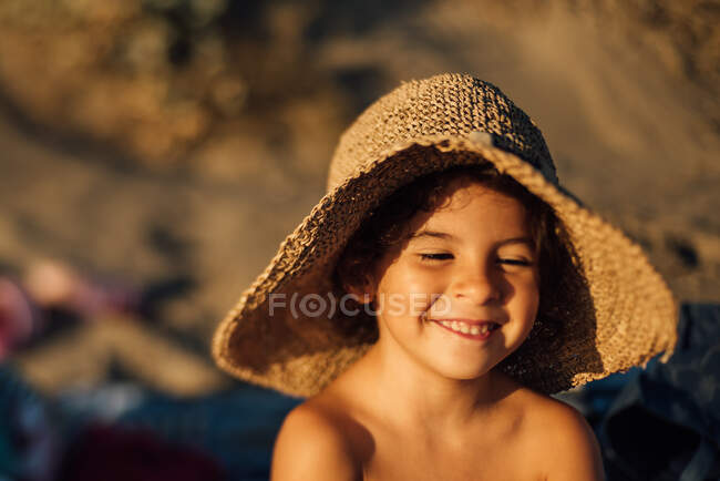 Ragazzina carina in cappello di paglia sorridente felicemente mentre riposa sulla spiaggia nella soleggiata giornata estiva — Foto stock