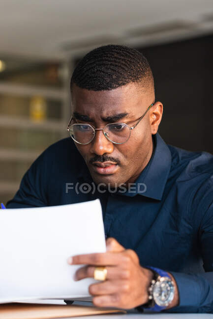Serious black adult male in formal clothes and eyeglasses sitting at table while taking notes in notebook from papers near netbook in bright workplace — Stock Photo