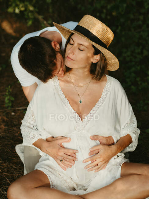 Мужчина обнимает и целует шею беременной женщины с закрытыми глазами, сидя на лугу на закате — стоковое фото