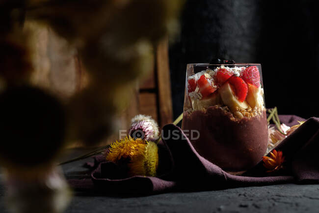 Tazón de batido apetitoso con fresas de granola y arándanos colocados en la mesa cerca de varias flores silvestres - foto de stock