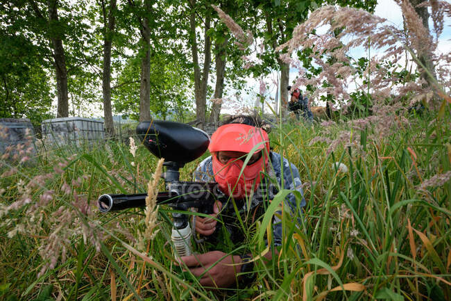 Неузнаваемый мужчина в шлеме и с пистолетом, нацеленным на противоположную команду во время игры в пейнтбол на природе — стоковое фото