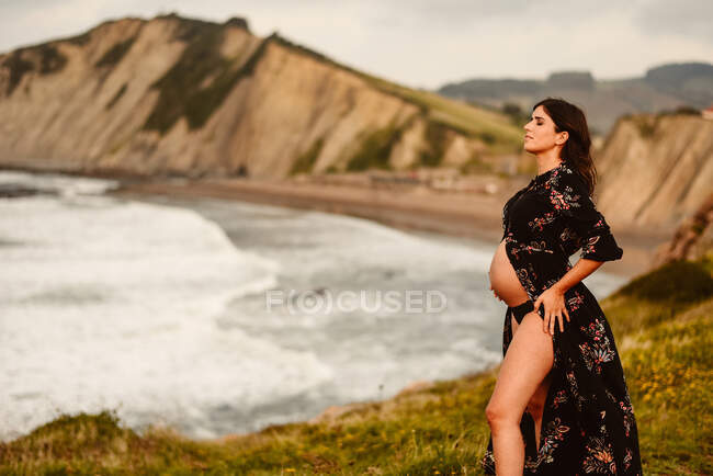 Vue latérale de paisible jeune hispanique femelle enceinte en robe élégante révélant ventre debout sur le bord de la mer contre les montagnes — Photo de stock