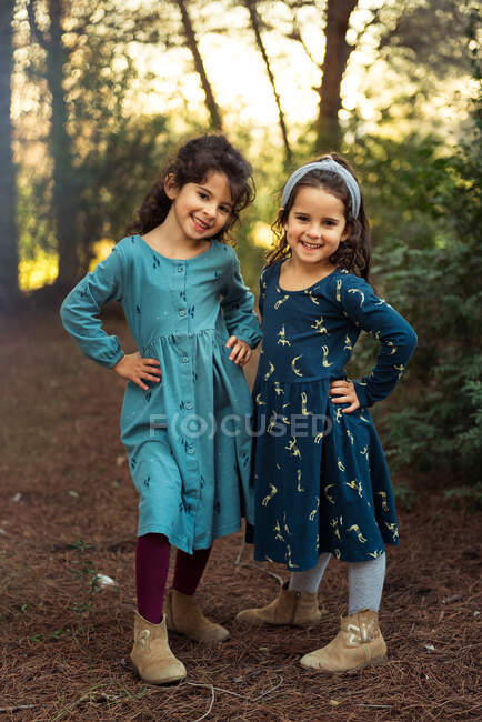Corpo inteiro de adoráveis irmãs sorridentes em vestidos olhando para a câmera enquanto estão juntos na floresta verde — Fotografia de Stock