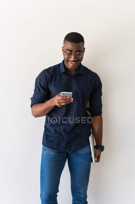 African American adult male in stylish outfit and eyeglasses standing with netbook in hands while using cellphone on white background — Stock Photo