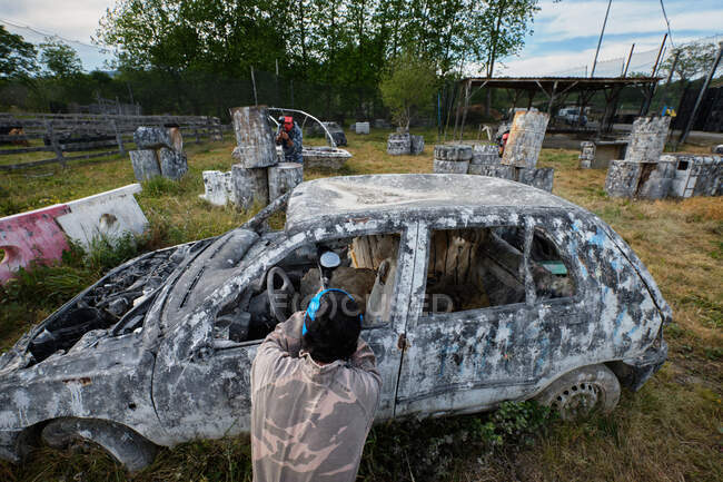 Hommes méconnaissables avec des armes et des casques de protection jouant au paintball parmi les voitures abandonnées — Photo de stock
