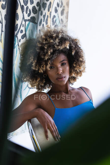 Moda hermosa mujer afroamericana con el pelo rizado y en vestido azul sentado en la silla en el estudio y mirando a la cámara - foto de stock