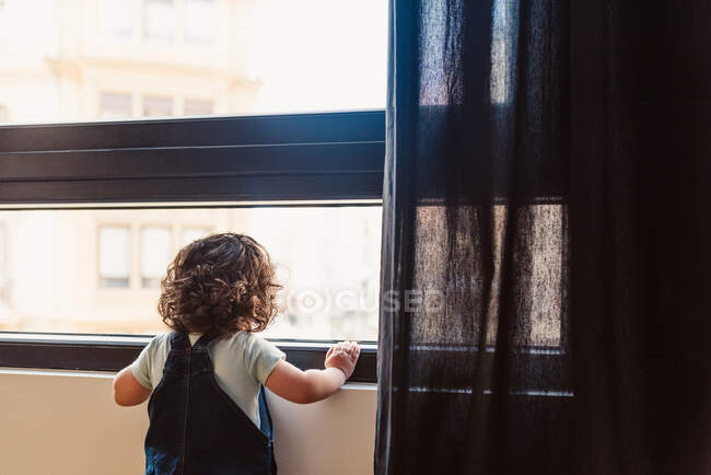 Vue de dos petit enfant méconnaissable avec des cheveux bouclés portant une combinaison en denim et regardant par la fenêtre tout en se tenant à la maison en plein jour — Photo de stock
