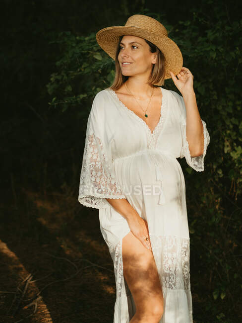 Serene pregnant female in dress and straw hat standing in countryside dark forest and looking away — Stock Photo