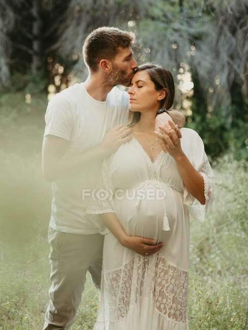 Нежный мужчина целует беременную женщину в лоб, стоя в поле на природе — стоковое фото