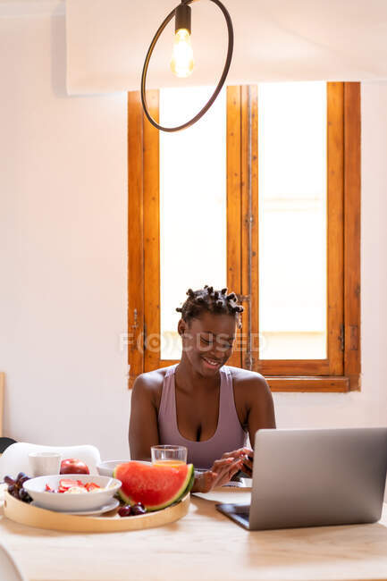 Smiling African American female browsing netbook while sitting at table with fresh fruits — Stock Photo