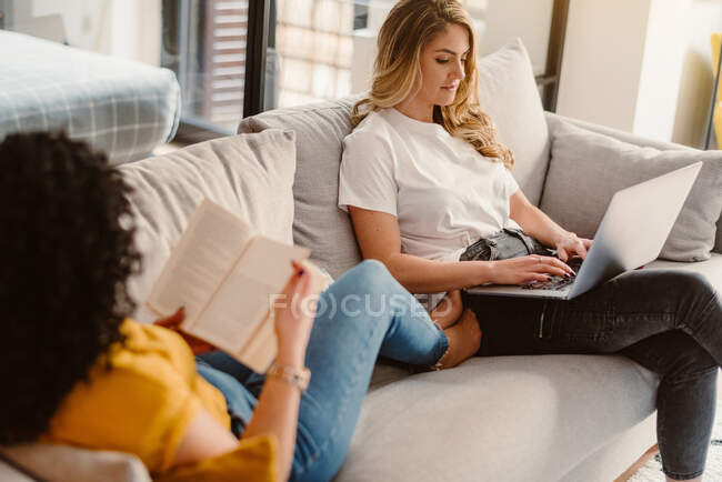 Pareja lesbiana navegando netbook y leyendo interesante libro mientras descansa en cómodo sofá en la sala de estar moderna - foto de stock