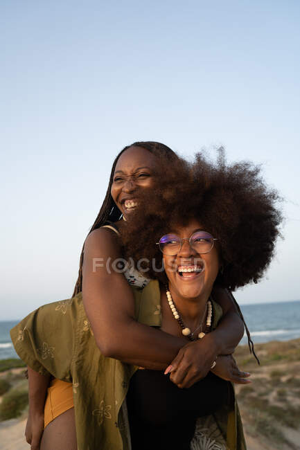Весела молода афроамериканка з афро - пігтеками їздить до смішної дівчини з кучерявим волоссям під час літніх канікул разом на березі моря. — стокове фото