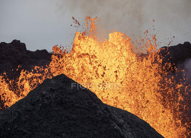 Salpicaduras de lava naranja caliente en erupción desde el pico de la montaña volcánica rodeado de humo en Islandia - foto de stock