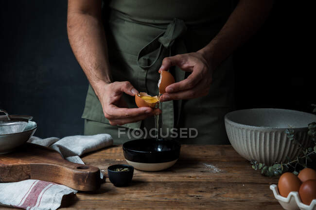 Chico irreconocible en delantal rompiendo huevo crudo sobre un tazón mientras prepara pastelería en una mesa de madera cerca de utensilios de cocina - foto de stock