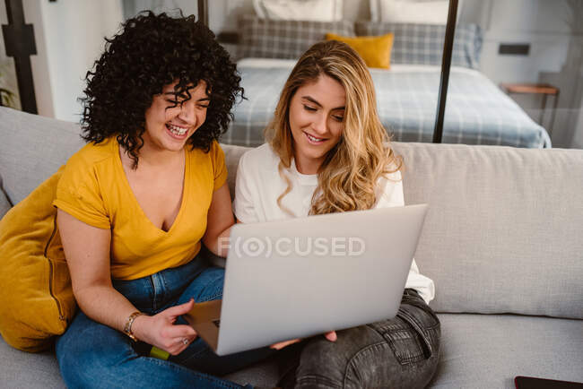 Joven pareja lesbiana navegando netbook juntos mientras están sentados en un acogedor sofá en la sala de estar - foto de stock