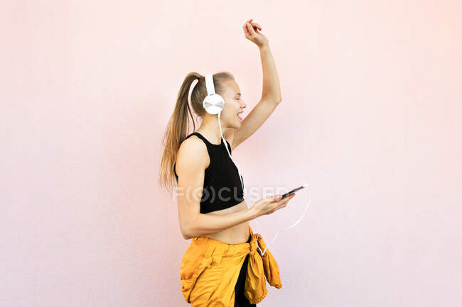 Jeune femme caucasienne portant un casque et une tenue de sport, écoutant de la musique au téléphone et dansant, isolée sur un fond lumineux — Photo de stock