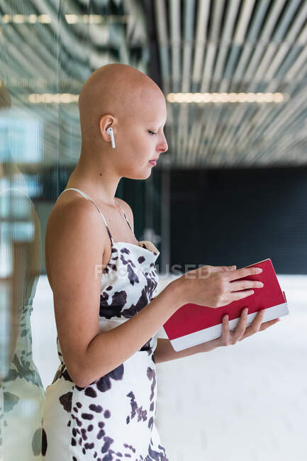 Бічний погляд на спокійну молоду лису жінку в стильному одязі і справжні бездротові навушники стоять на вулиці біля будівлі зі скляними стінами і читають цікаву книгу — стокове фото