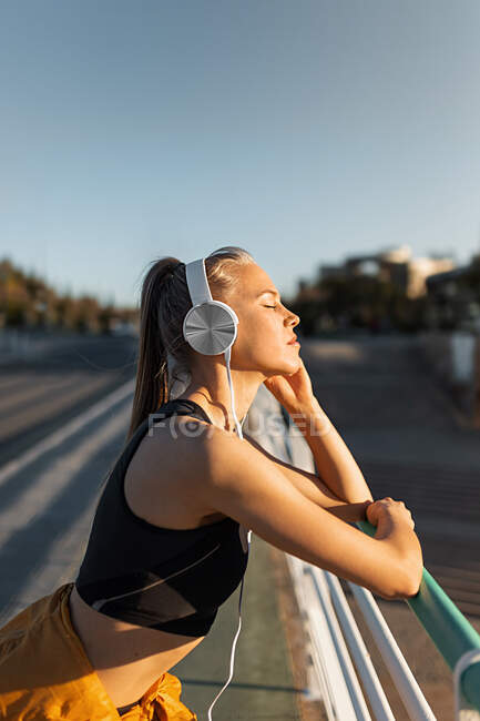 Young athletic caucasian leaning on banister outdoors, listening to music and relaxing with closed eyes after working out — Stock Photo