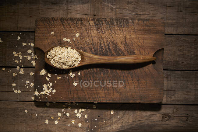 Composición de vista superior con cuchara de madera llena de copos de avena saludables colocados en la tabla de cortar en la mesa de tablón rústico - foto de stock